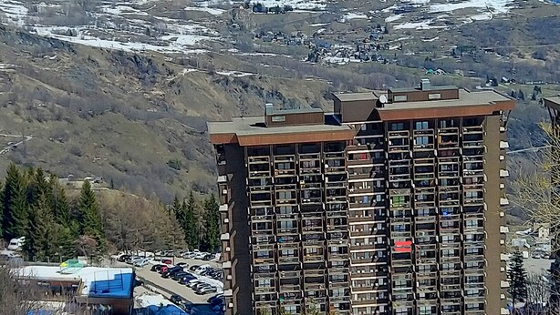 Un edificio de gran altura con múltiples balcones, rodeado de montañas nevadas y un estacionamiento.