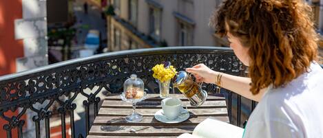 A woman reading a book at a wooden table with a cup and saucer, a glass of tea, and a vase of flowers on a balcony.