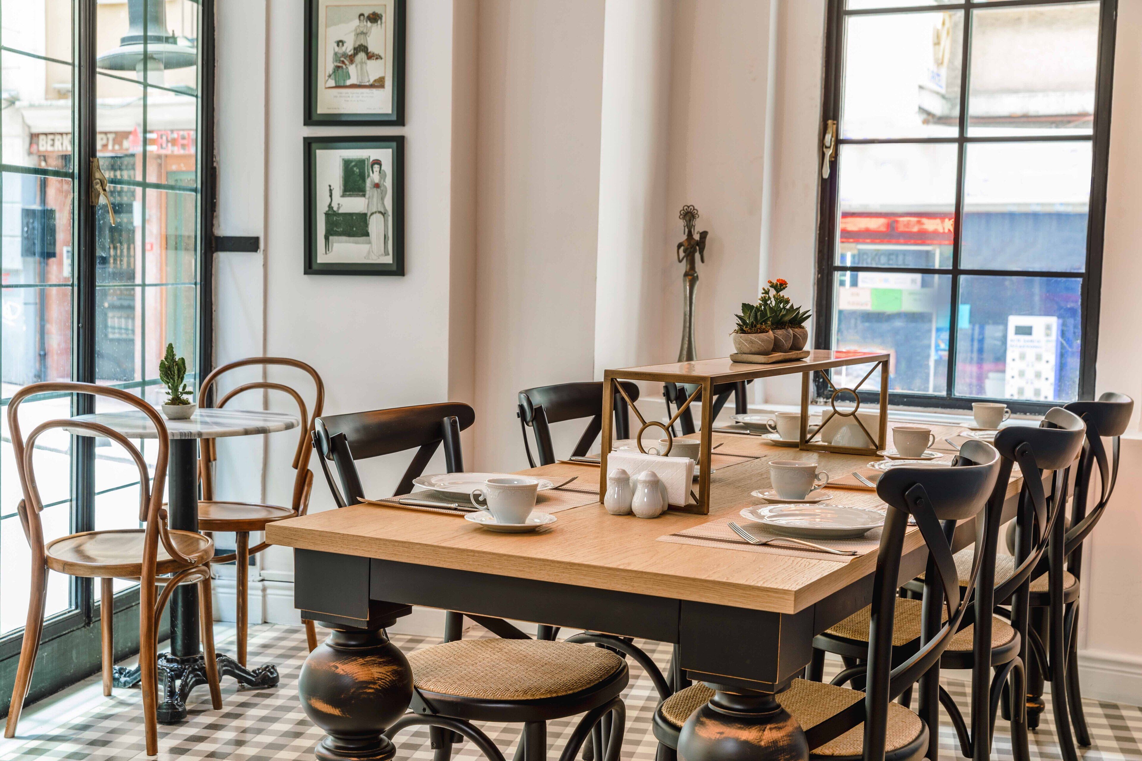 Un coin repas avec un sol à carreaux, des tables et des chaises en bois. Les murs sont ornés de tableaux encadrés et une fenêtre offre une vue sur les bâtiments à l’extérieur.