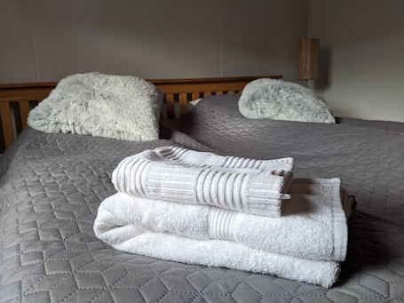 A neatly made bed with a quilted grey bedspread, two fluffy white pillows, and a folded white towel.. Engimýri Lodge