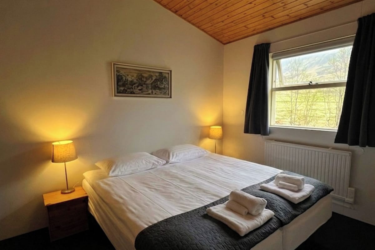A bedroom with a wooden ceiling, a large bed with two pillows, a window with curtains, and two bedside lamps.