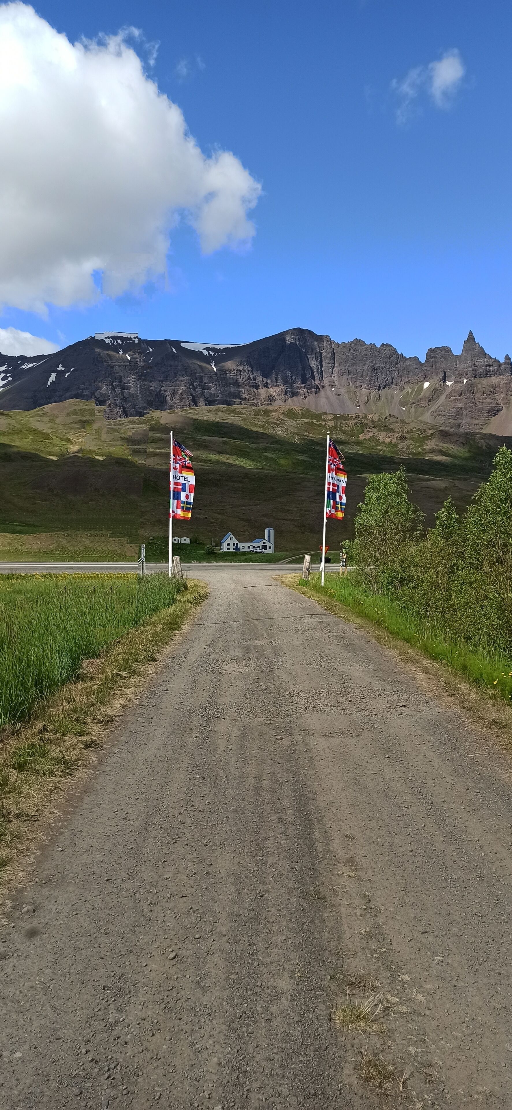 A dirt road leading to a building with a flag in the foreground and a mountain in the background.