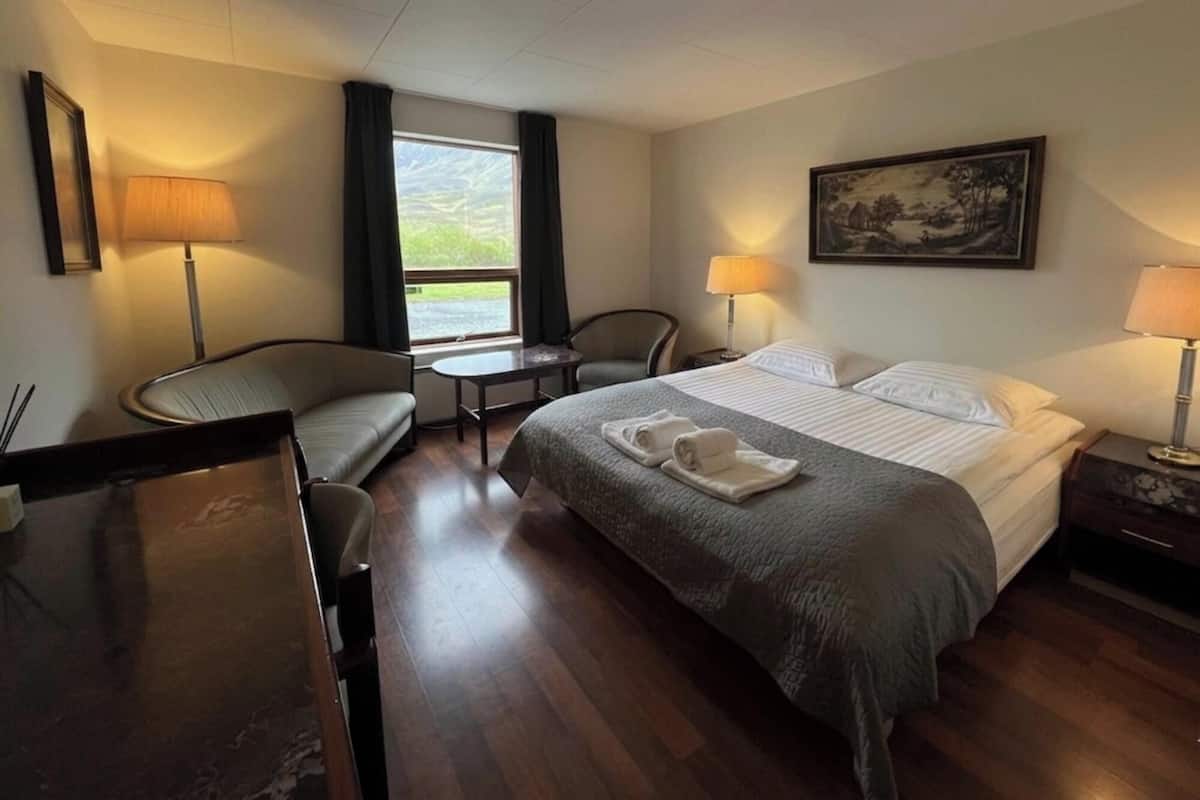 Habitación de hotel con cama, dos sillas, una mesita, ventana con cortinas, un cuadro en la pared y una lámpara a cada lado de la cama.