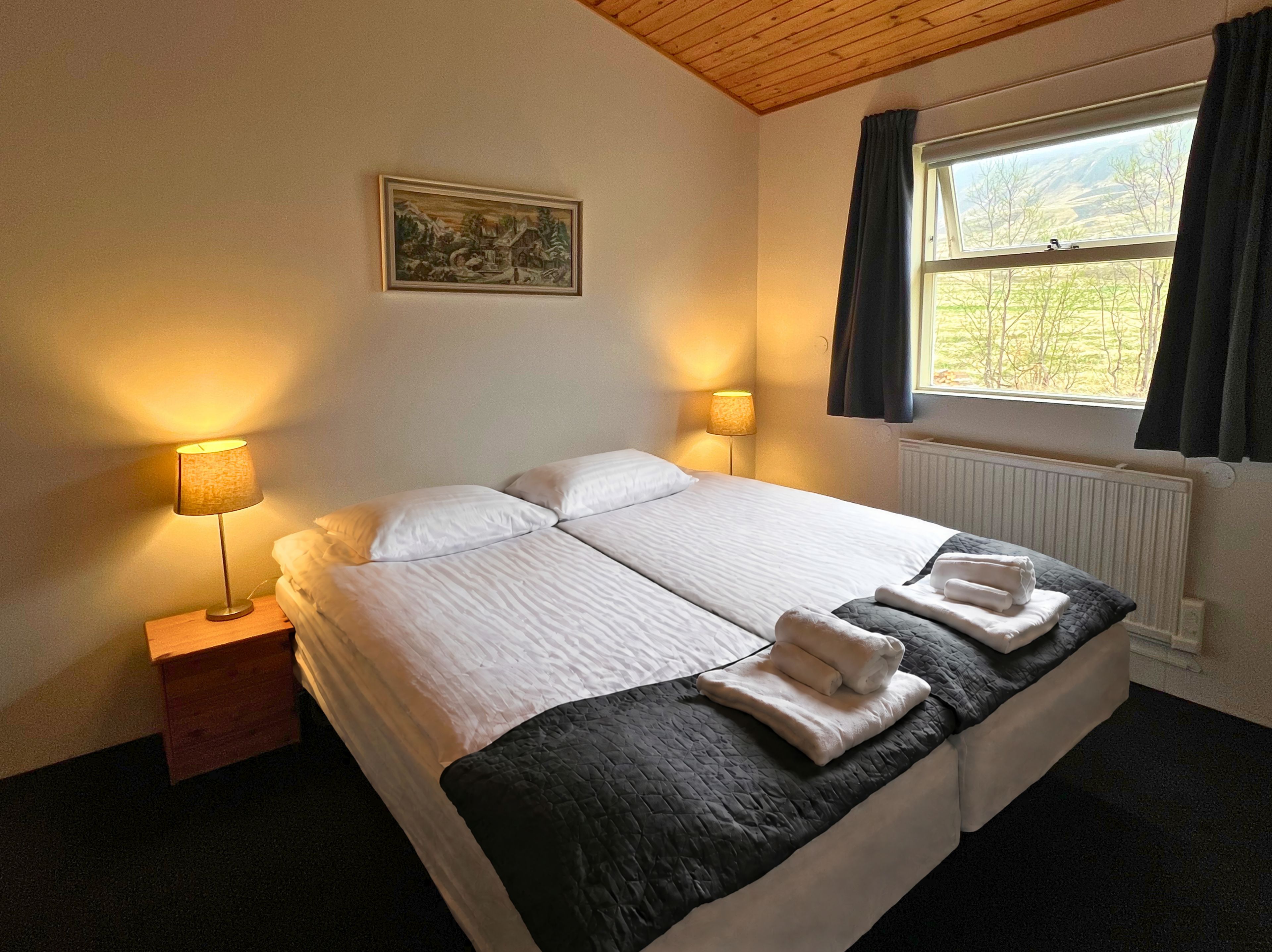 Una cama doble con dos almohadas, una mesita de noche con lámpara, una ventana con cortinas y un cuadro en la pared.