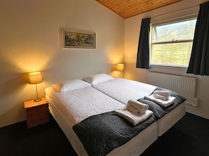 A double bed with two pillows, a bedside table with a lamp, a window with curtains, and a painting on the wall. - Engimýri Lodge (Bakki)
