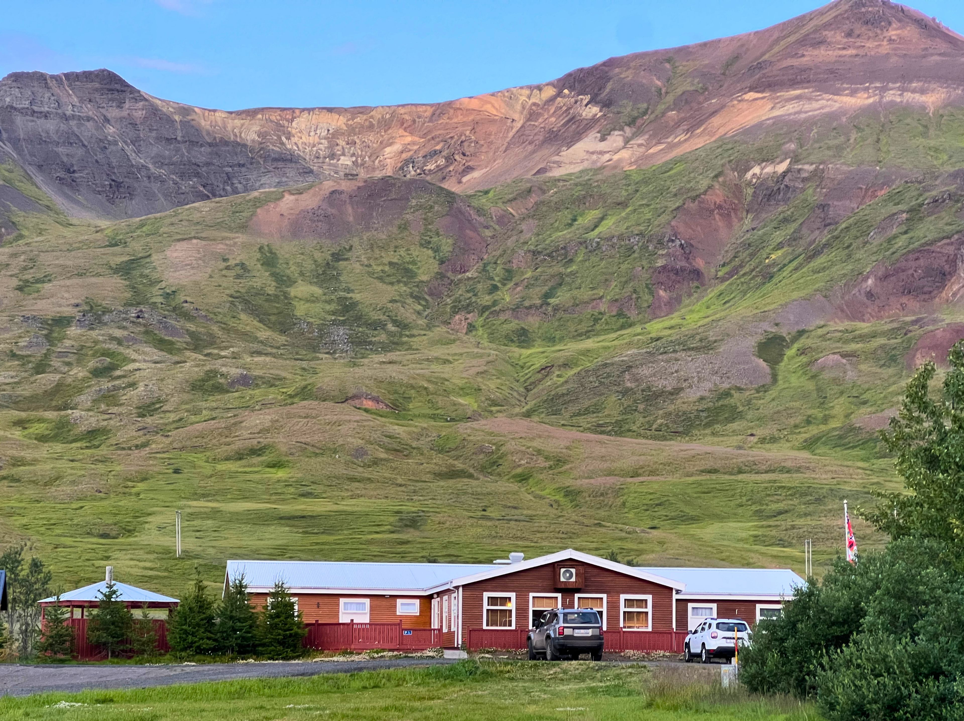 Un edificio rojo con techo plano, dos coches estacionados adelante y un paisaje montañoso al fondo.