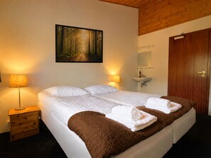 A double bed with a brown quilt, two bedside lamps, a painting of a forest, a wooden door, and a small bedside table. - Engimýri Lodge (Bakki)