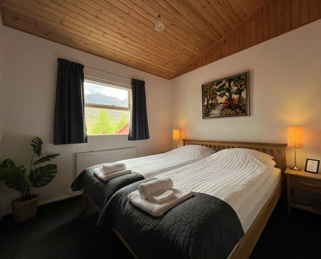 A room with a wooden ceiling, a bed with two pillows, a window with curtains, a plant, and a painting on the wall.