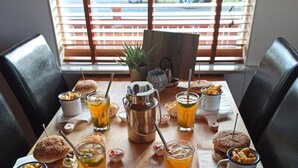 A dining table with food and drinks, a metal coffee pot, and a view of the outside through a window with blinds. - Engimýri Lodge (Bakki)