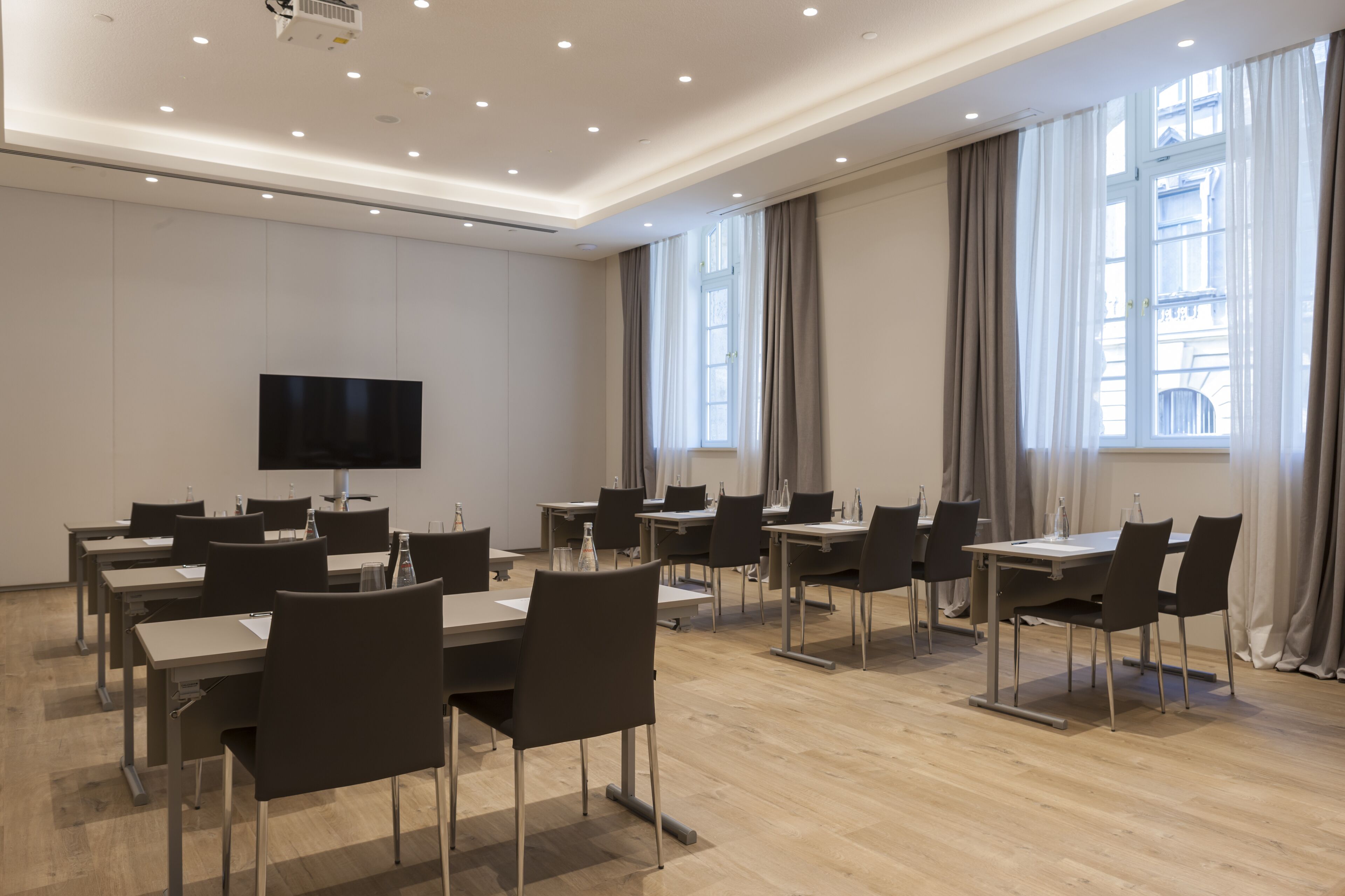Un salón de conferencias con mesas y sillas, un televisor de pantalla plana y ventanas grandes con cortinas.
