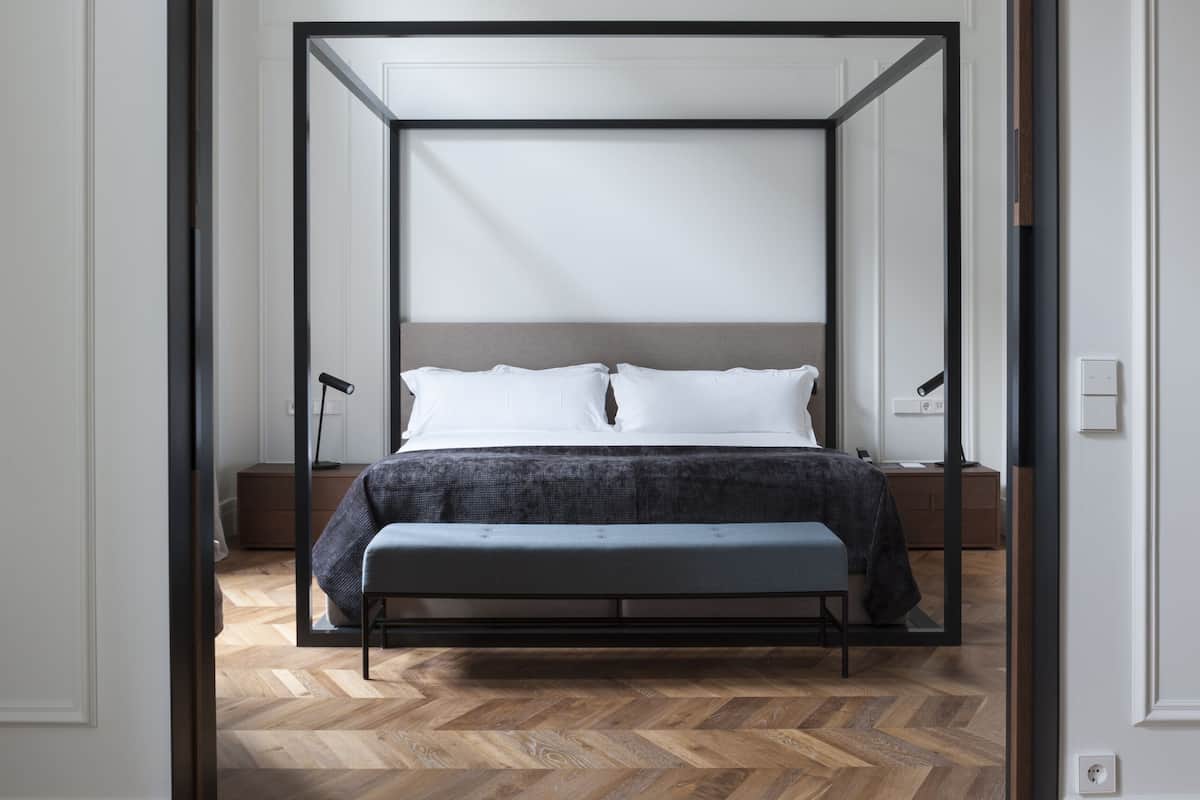 A bedroom with a four-poster bed, a wooden herringbone floor, and a white door frame.