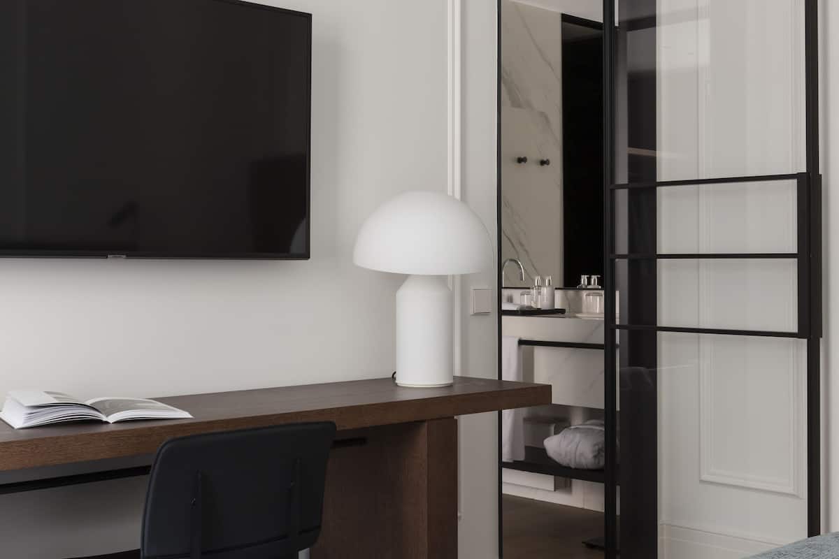 A modern hotel room with a wooden desk, a black chair, a flat-screen TV, a white lamp, and a bed with a grey blanket.
