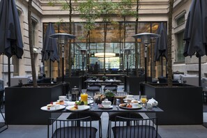 Un espacio de comedor al aire libre con mesas, sillas, sombrillas y vistas a un edificio a través de paredes de vidrio.