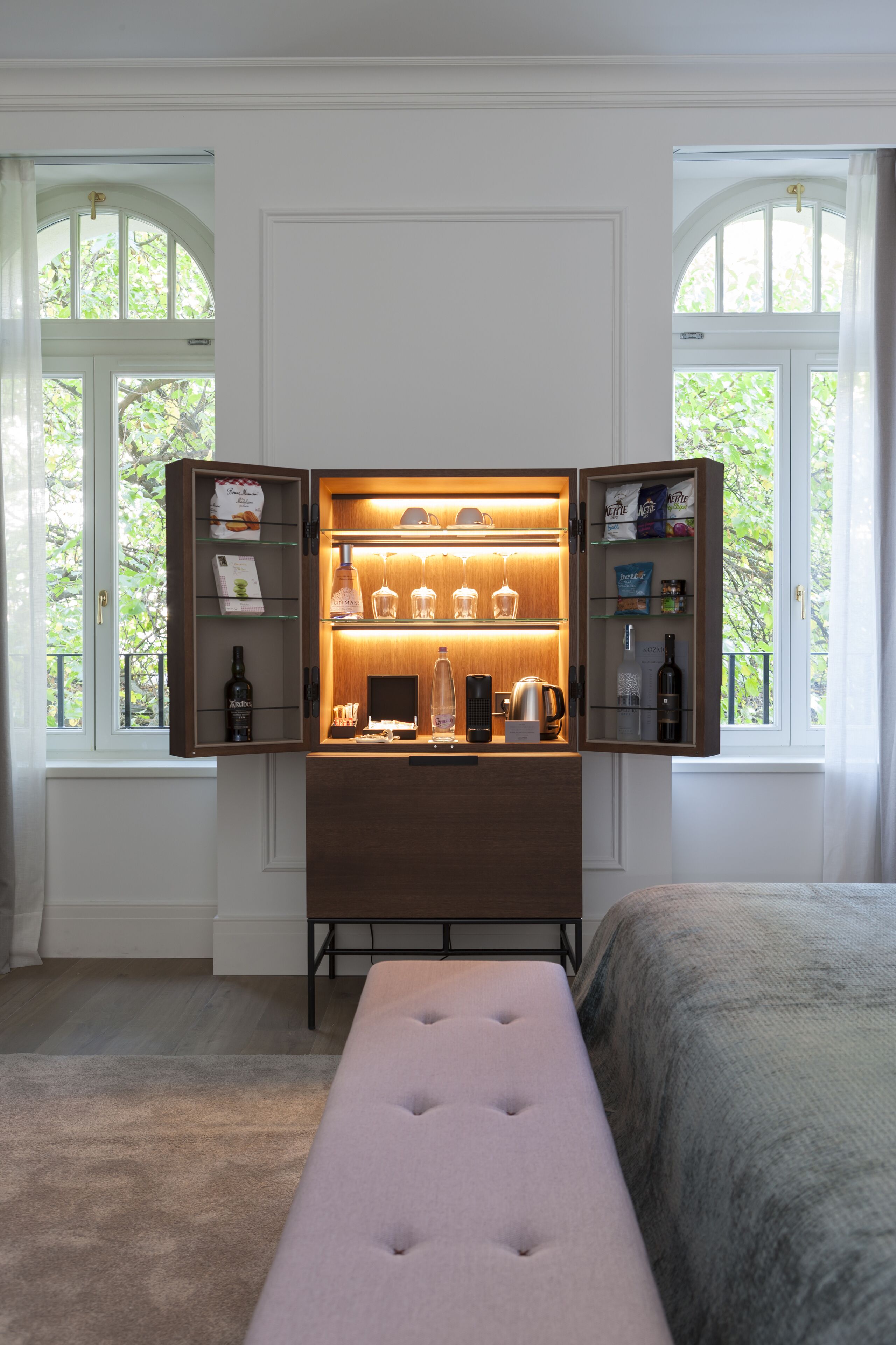 Ein Zimmer mit einem Bett, einer Sitzbank, einer Minibar mit Gläsern und Blick auf Bäume durch Bogenfenster.