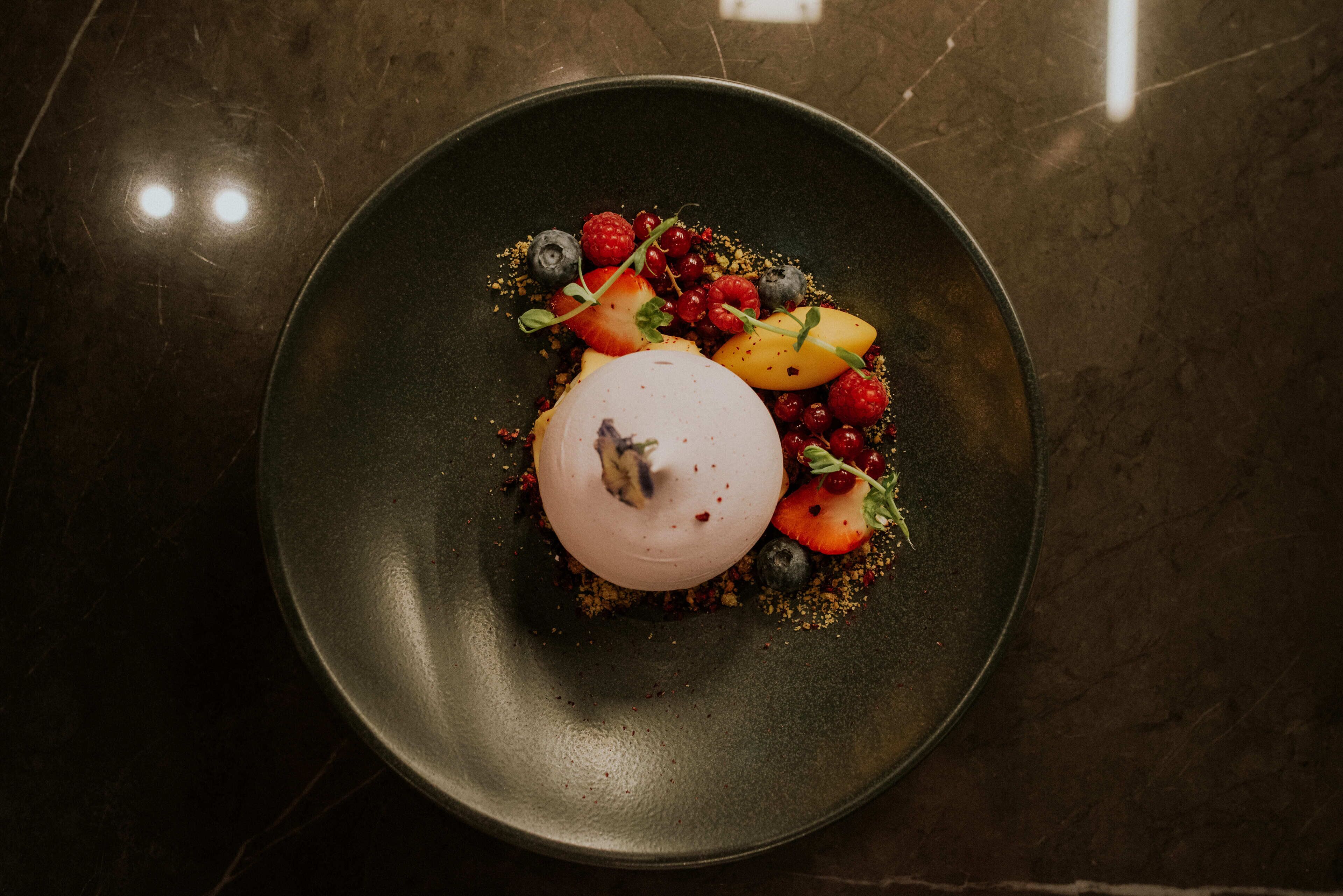 Un postre en un plato oscuro con varias frutas y bayas.