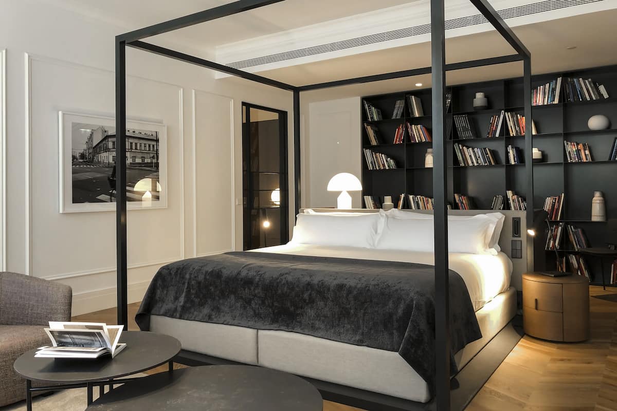 A modern bedroom with a four-poster bed, a round coffee table, a bookshelf, and a framed picture on the wall.