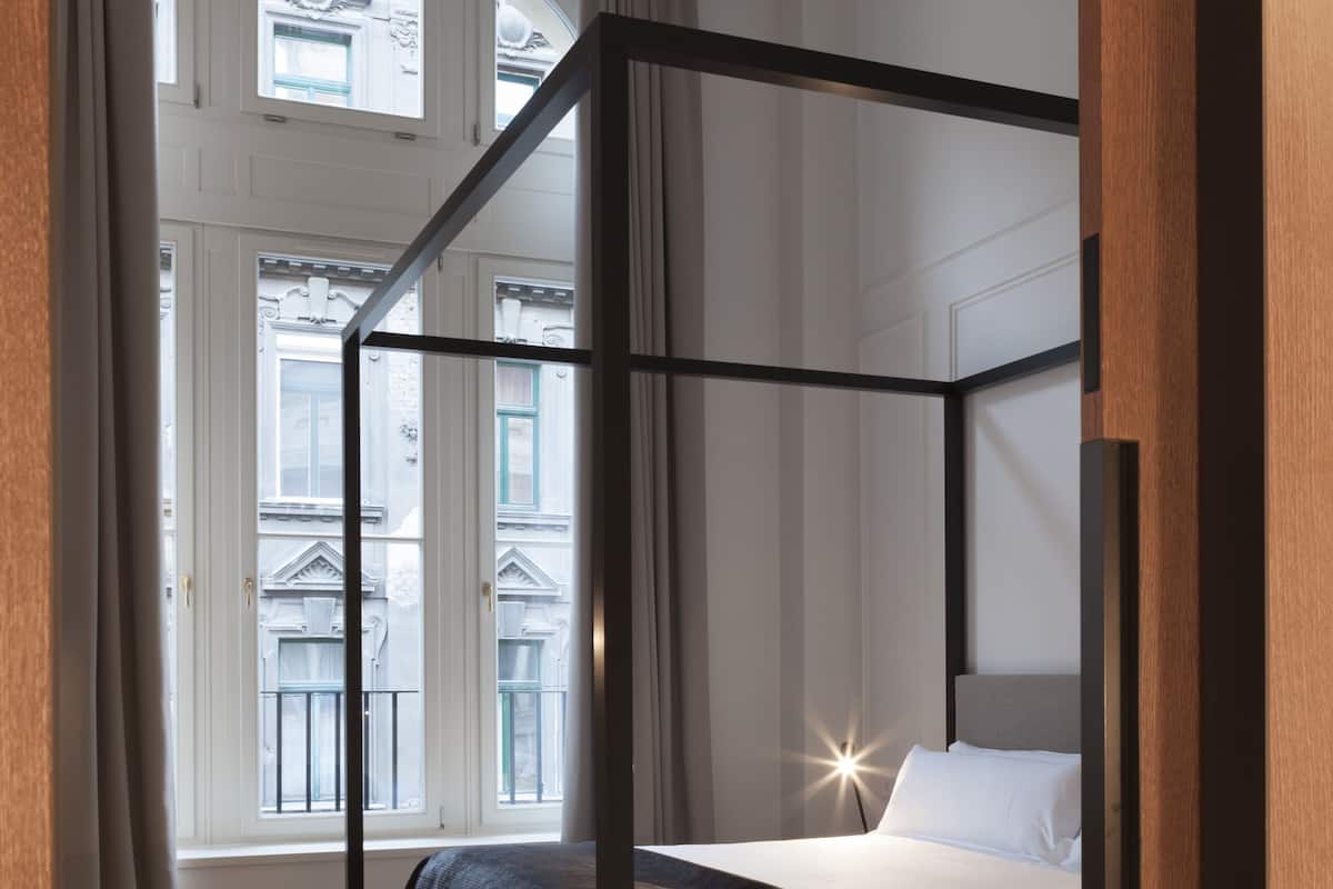 A four-poster bed with a bench, a bedside table, and a view of buildings through large windows.