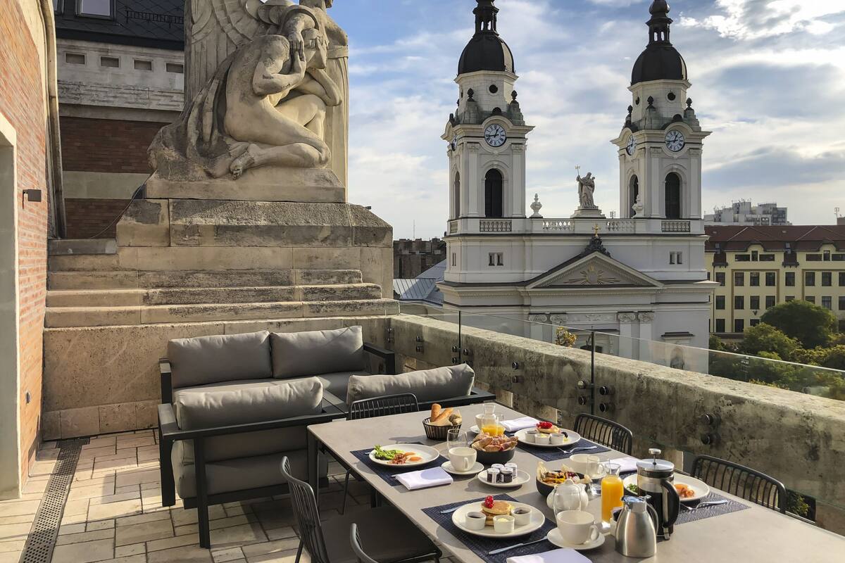 A rooftop terrace with a dining table set for breakfast, a statue, and two clock towers in the background.