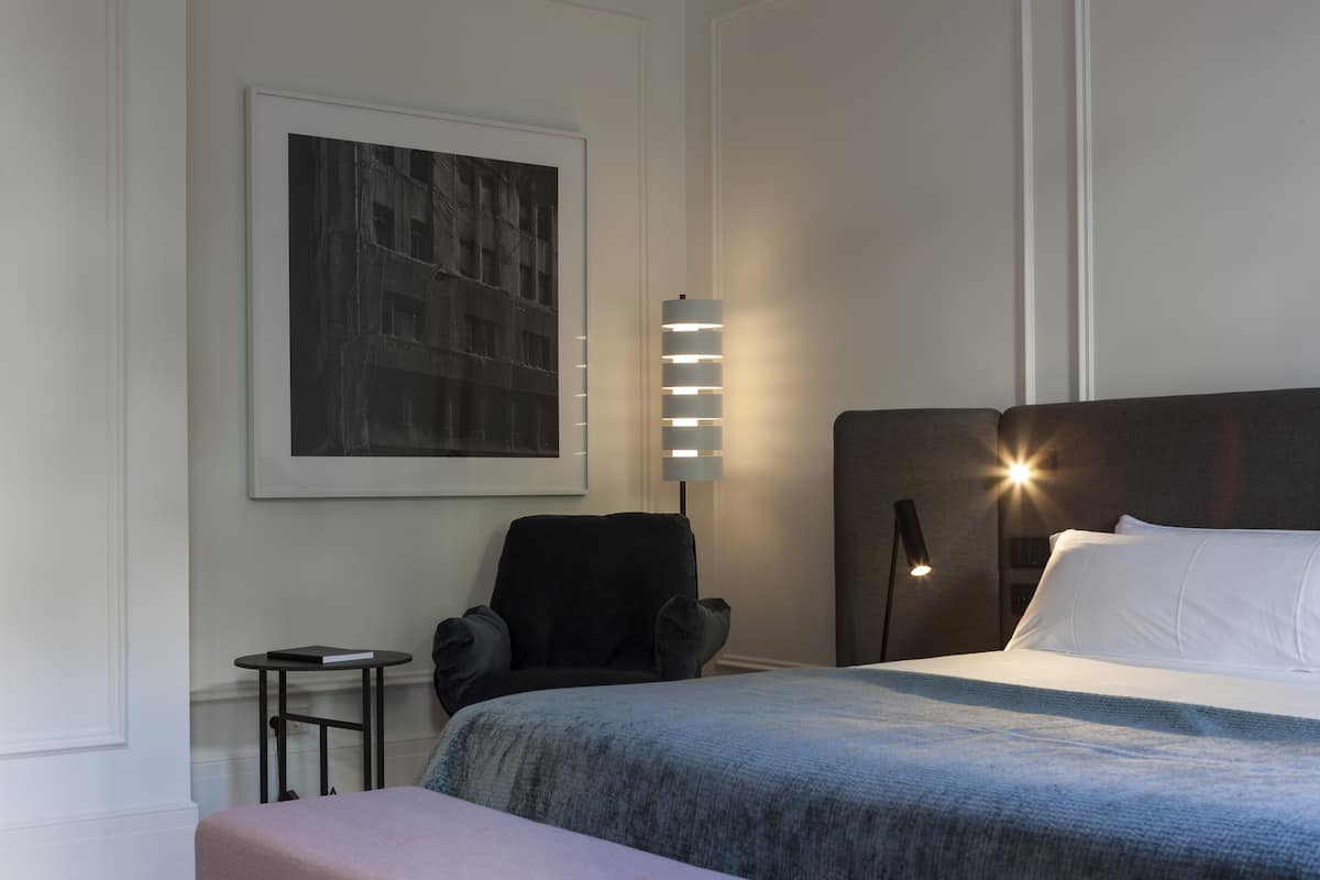 A hotel room with a bed, a chair, a small table, and a wall-mounted lamp.