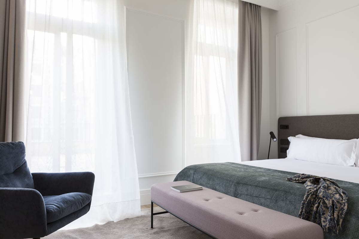 A hotel room with a bed, a bench, a chair, and sheer curtains.