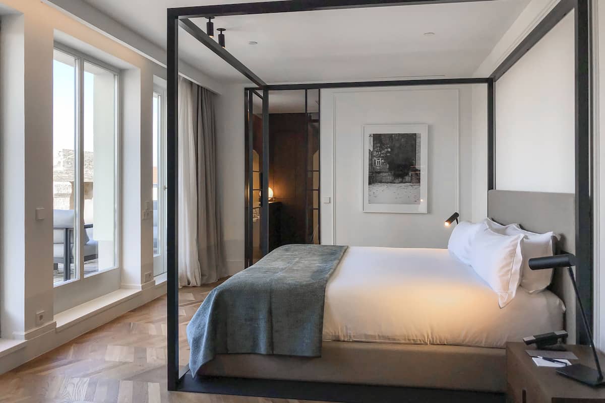 A modern hotel room with a large bed, a bedside table, a lamp, and a view of the outdoors through large windows.