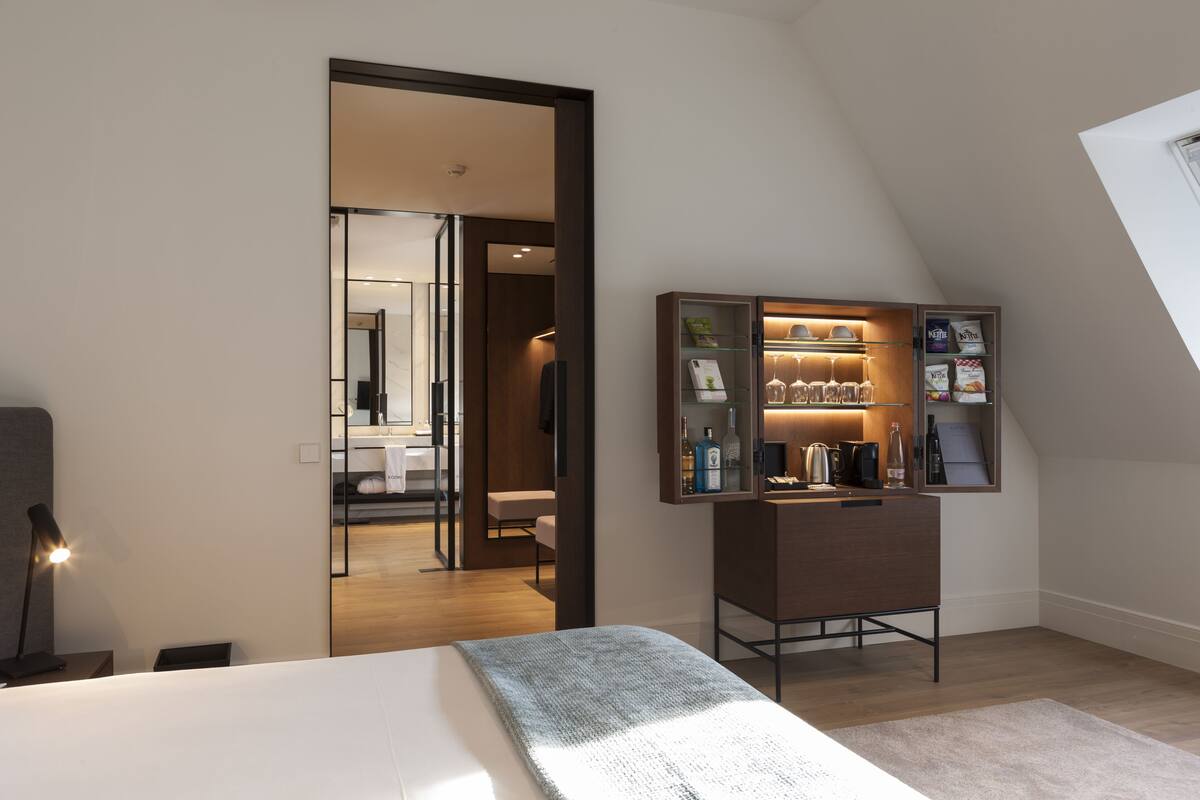 A modern hotel room with a bed, a bedside lamp, a small cabinet with snacks, and a view of the adjacent dining area.