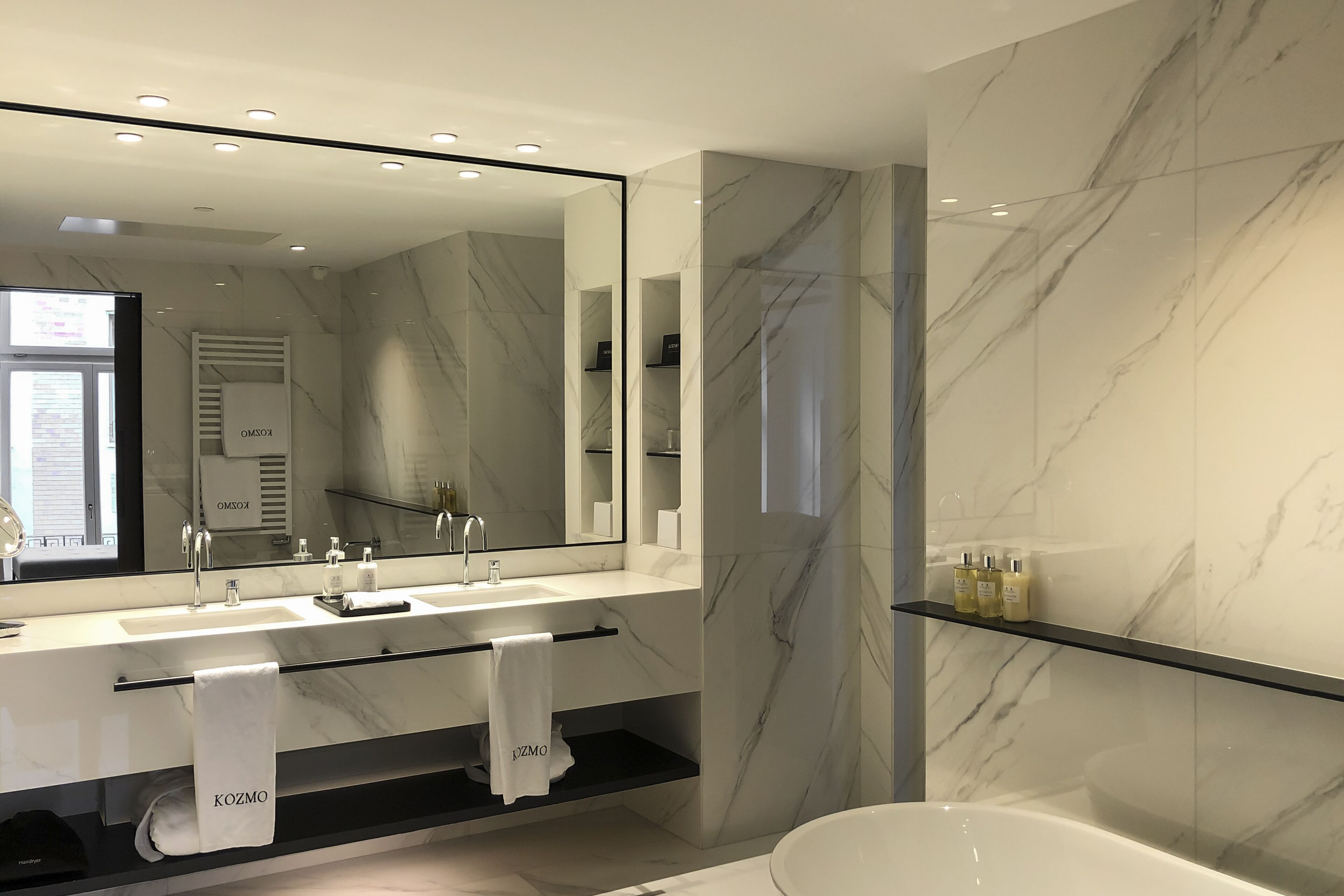 A modern bathroom with a large mirror, two sinks, a bathtub, and marble walls.