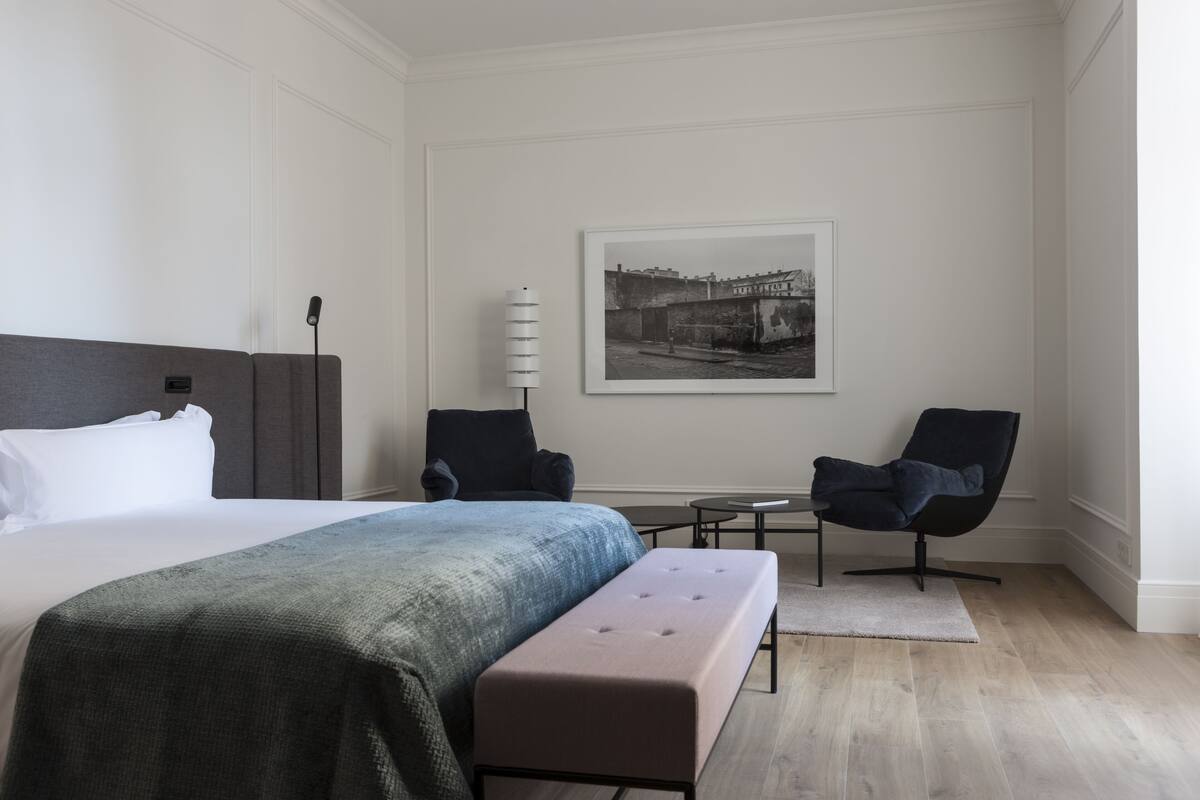 A modern hotel room with a large bed, a bench, two armchairs, a small table, and a framed picture on the wall.