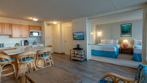 A compact hotel room with a kitchenette, a dining area with wicker chairs, a bed with blue bedding, and a painting of a beach scene. - Block to Beach! Koko Resorts at the Waikiki Banyan 22nd Floor Pool View, Free parking & Wifi (Honolulu)