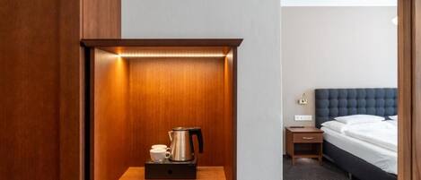 Habitación de hotel con un armario de madera, una mesita de noche con hervidor, una taza y un cajón pequeño.