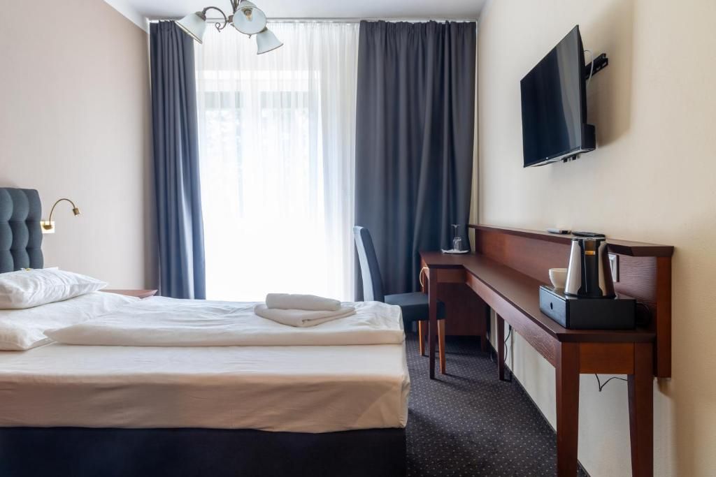 Ett hotellrum med en säng, ett skrivbord, en stol, en tv och ett fönster med gardiner.