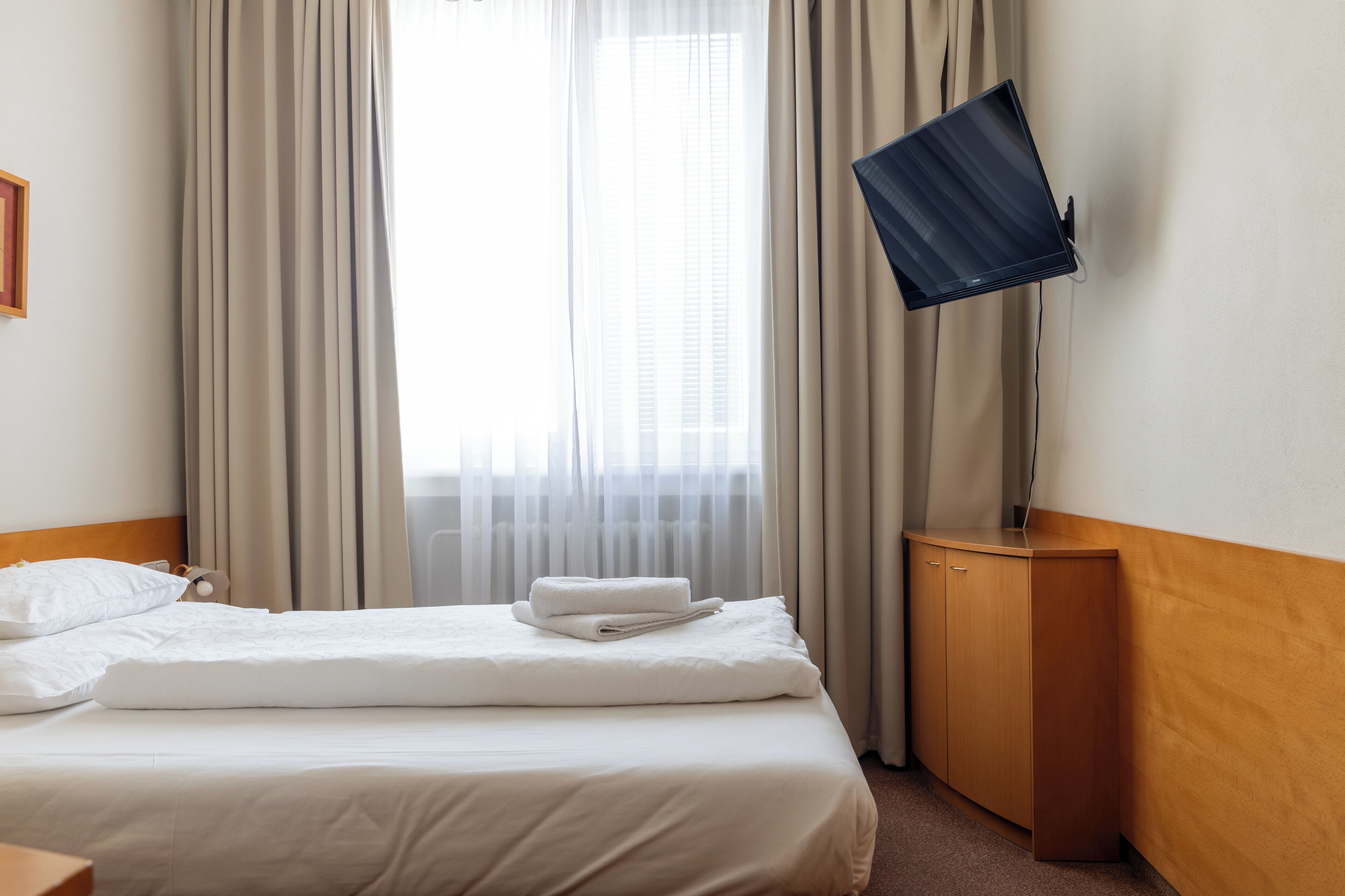 A hotel room with a bed, a TV mounted on a stand, and a window with curtains.