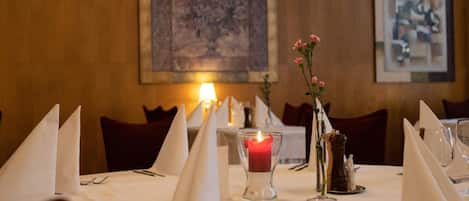 Una mesa de restaurante con manteles blancos, cubiertos, una vela encendida y un florero con flores rosadas.