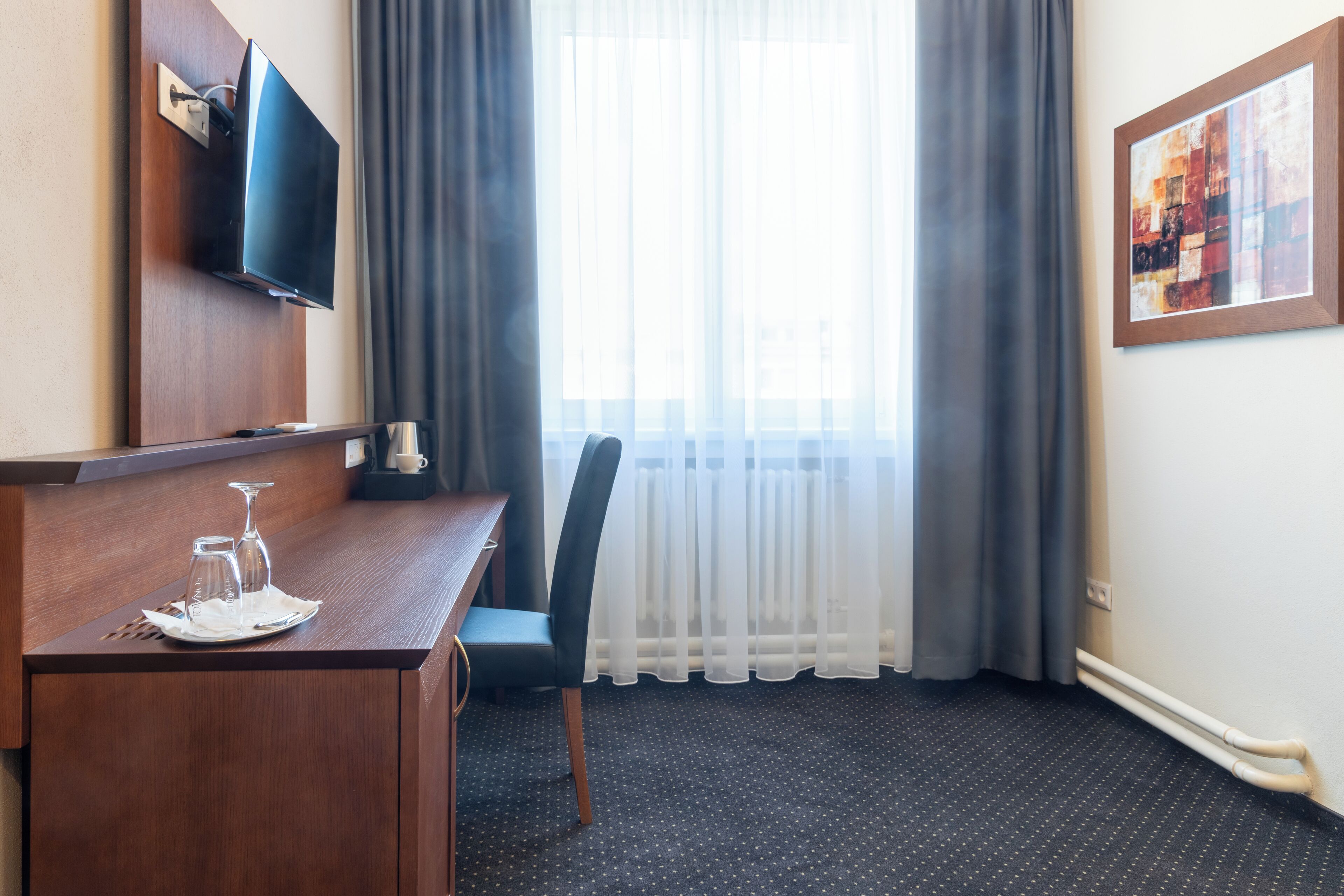 A hotel room with a wooden desk, a flat-screen TV mounted on the wall, a chair, a window with sheer curtains, and a framed artwork on the wall.