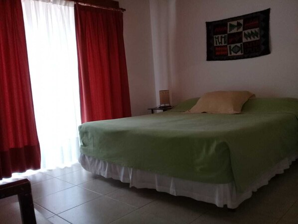 Un dormitorio con cama, cortinas rojas, un tapiz en la pared y suelo de baldosas.