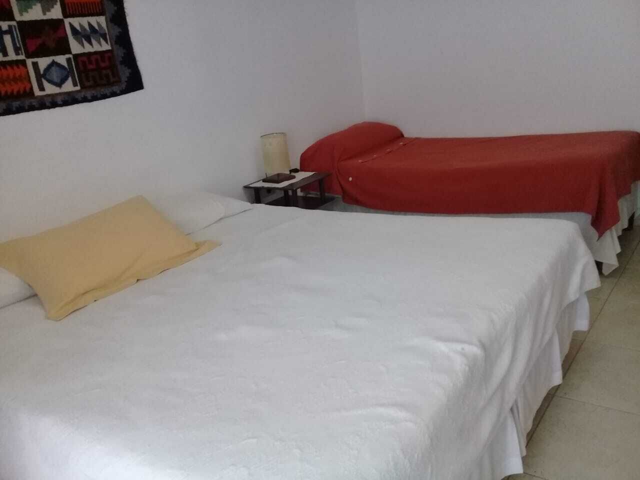 Una habitación con dos camas, una con almohada amarilla y la otra con una colcha roja. Hay una mesita con una lámpara y un teléfono.