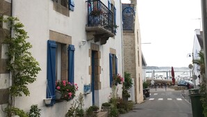 A narrow street with a white building featuring blue shutters and a balcony with flowers. - Le Grand Large - house in Locmariaquer centre, 4p (Locmariaquer)