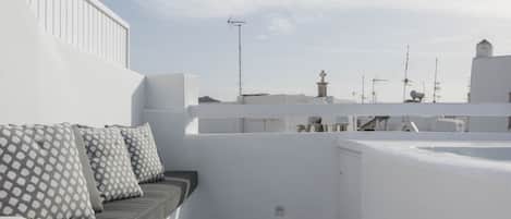 Una terrazza sul tetto con un muro bianco, una panca imbottita con cuscini decorati e due tavolini con gambe in metallo.