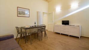 Een kamer met een eettafel, stoelen, een bank, een tv op een kast en een lijst met een foto aan de muur.