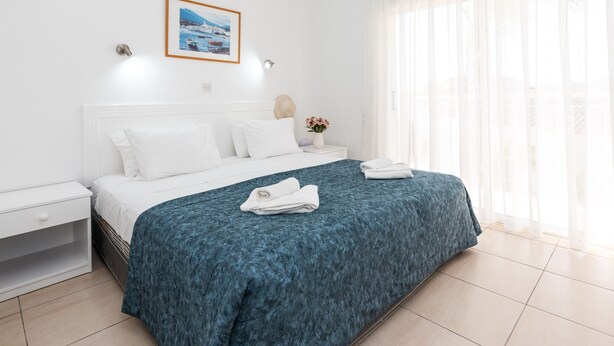Ein ordentlich bezogenes Bett mit weißen Laken und einer blauen, strukturierten Tagesdecke, ein weißer Nachttisch mit Schublade, ein gerahmtes Bild an der Wand und ein Fenster mit durchsichtigen Vorhängen.