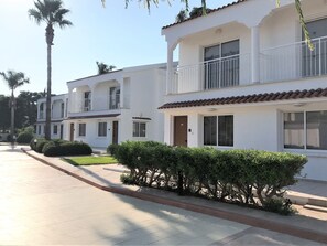 Une rangée de bâtiments blancs à deux étages, dotés de balcons, avec un trottoir pavé et des palmiers.