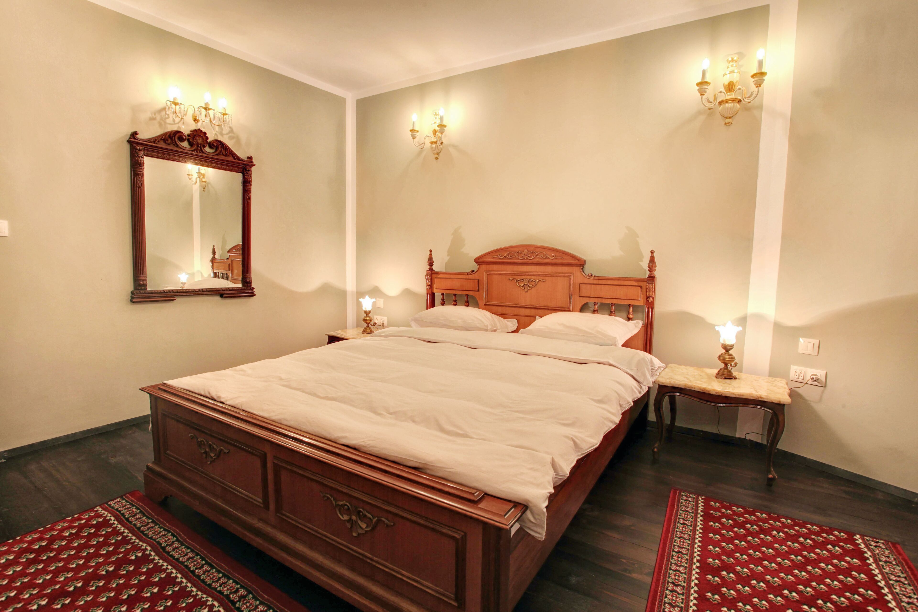 Una cama bien hecha con cabecera de madera, una mesita de noche con lámpara y un espejo grande en la pared.