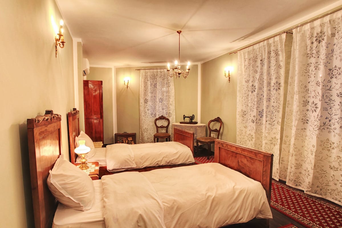 A hotel room with two beds, a wooden wardrobe, a mirror, a chandelier, and floral curtains.