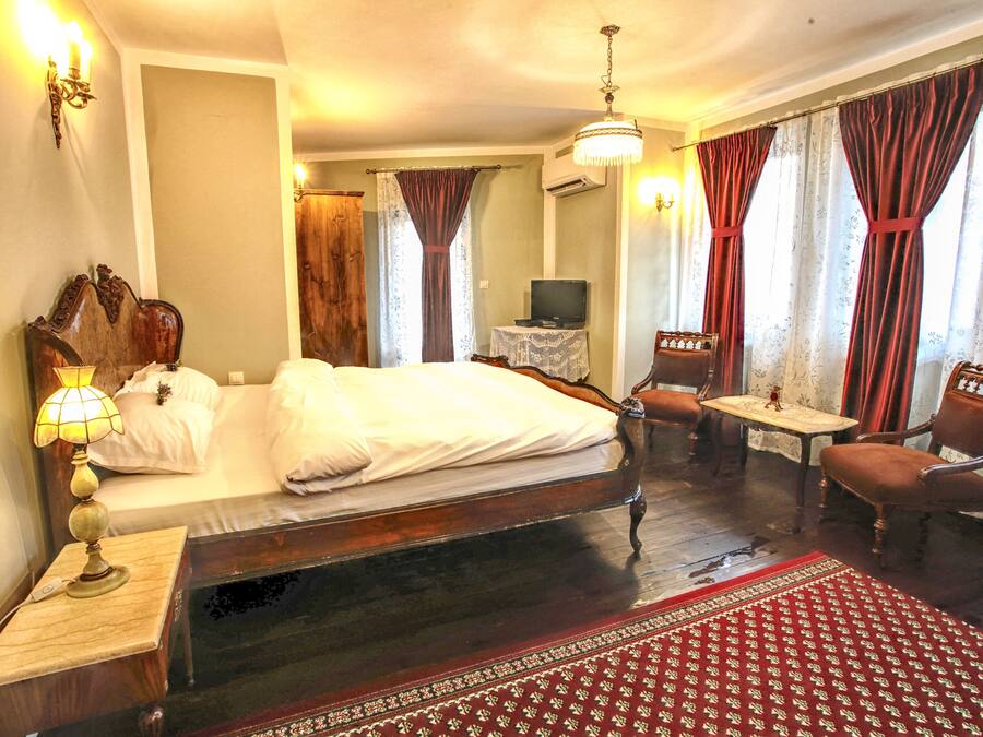 Habitación de hotel con una cama grande, una mesita de noche de madera, una mesa con televisor, una silla y un tapete rojo estampado.