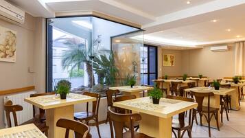Un espace de restauration moderne avec des tables et des chaises en bois, des plantes en pot et de grandes fenêtres offrant une vue sur les palmiers et les bâtiments.