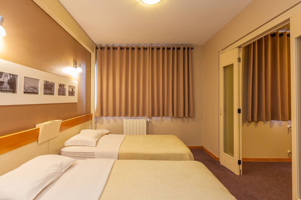 Ein Hotelzimmer mit zwei Betten, einem Heizkörper, einer Wandleuchte und einer Tür mit Vorhängen.
