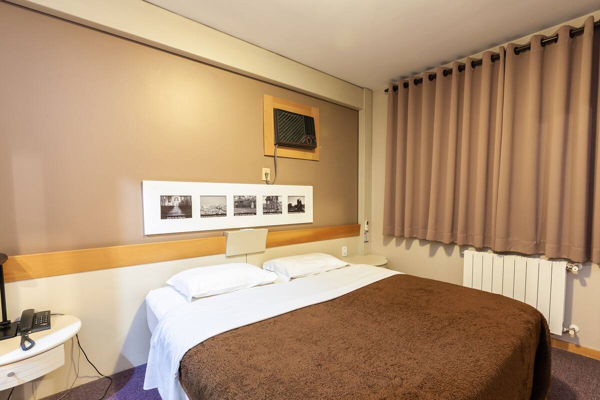 Ein Hotelzimmer mit Bett, Nachttisch mit Lampe, Wandklimaanlage und einem Fenster mit Vorhängen.