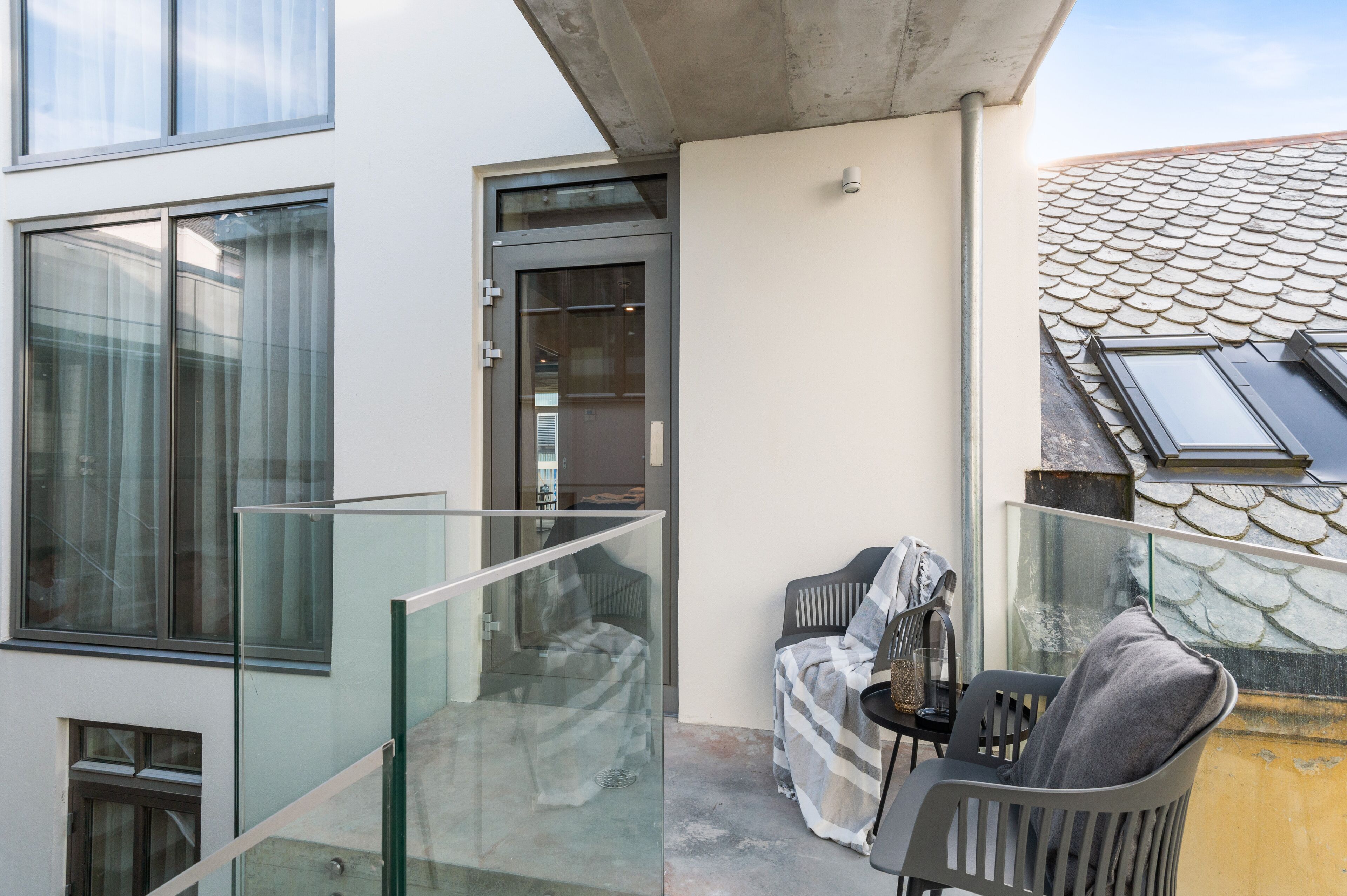 A modern apartment with a glass balcony, a sliding glass door, and a view of a tiled roof.