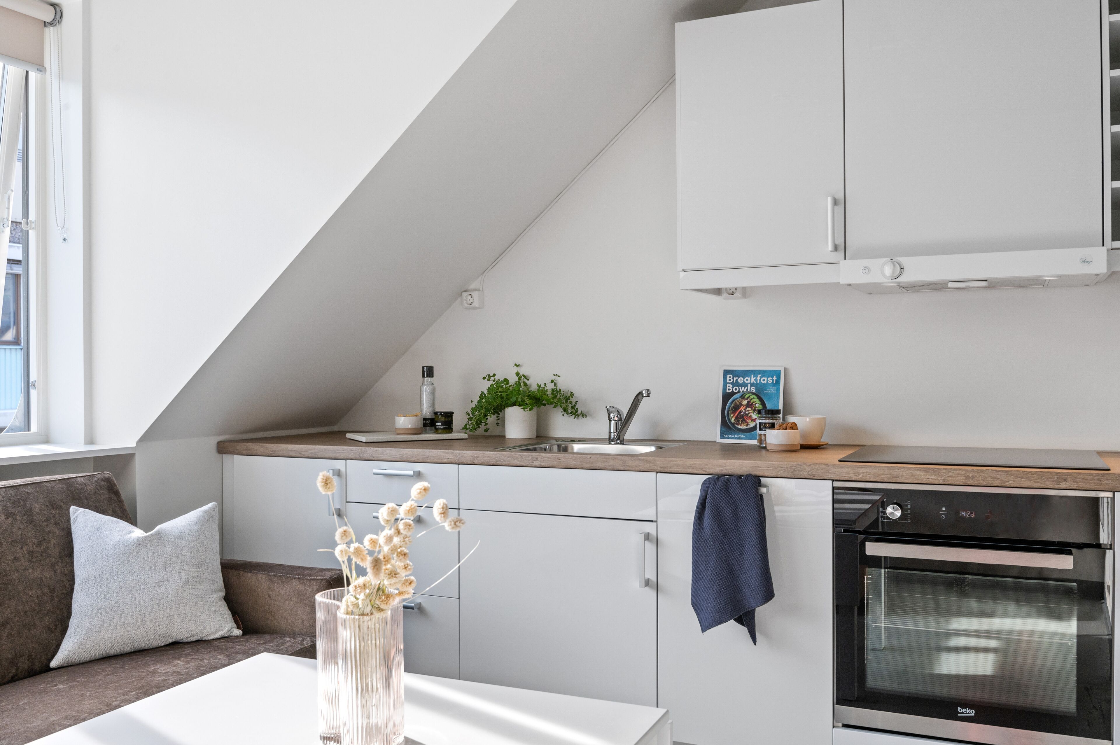 Una cocina moderna con armarios blancos, horno integrado y comedor con mesa y sofá.