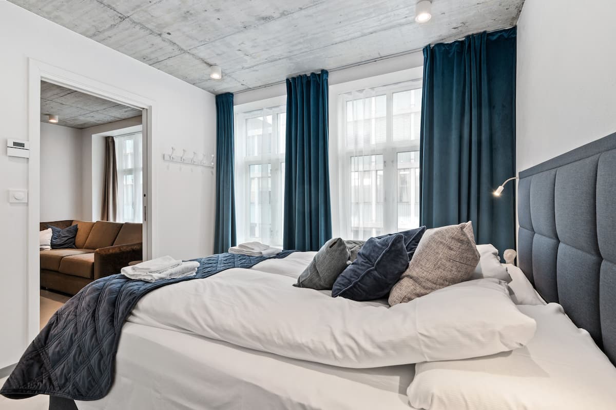 Une chambre moderne avec un grand lit, une tête de lit rembourrée de couleur grise et une vue sur le paysage urbain, grâce à de grandes fenêtres ornées de rideaux bleus.